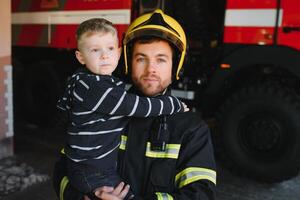 een brandweerman nemen een weinig kind jongen naar opslaan hem. brand motor auto Aan achtergrond. brandweerman met kind in zijn armen. bescherming concept. foto