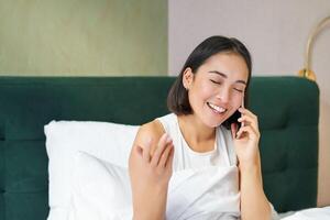 dichtbij omhoog portret van schattig Aziatisch meisje in bed, pratend Aan mobiel telefoon met gelukkig glimlachen gezicht. vrouw ontwaken omhoog en maken een telefoon telefoontje foto