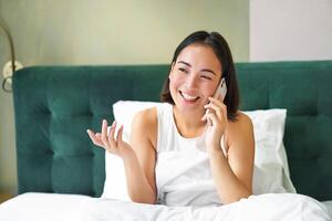 vrouw praat Aan mobiel telefoon in bed. glimlachen meisje hebben telefoon gesprek terwijl ontspannende in slaapkamer foto