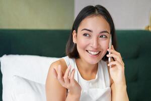 dichtbij omhoog portret van schattig Aziatisch meisje in bed, pratend Aan mobiel telefoon met gelukkig glimlachen gezicht. vrouw ontwaken omhoog en maken een telefoon telefoontje foto