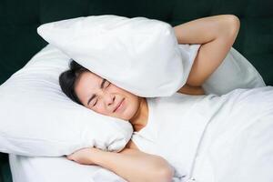 verdrietig meisje met slapeloosheid. jong Aziatisch vrouw aan het liegen in bed, Hoes haar oren met kussen, kan niet slaap, partner snurkt, luid lawaai Bij nacht verontrustend haar foto