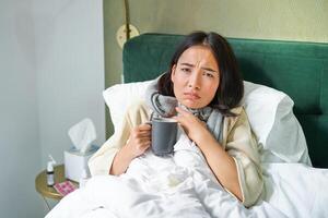 Gezondheid, griep en mensen concept. portret van Koreaans vrouw gevoel ziek, vangst een koud, blijven in bed onwel, drinken heet thee foto