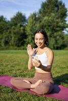 portret van Aziatisch meisje zittend Aan mat in park, pratend naar smartphone, mediteren online met yoga instructeur Aan vers lucht foto