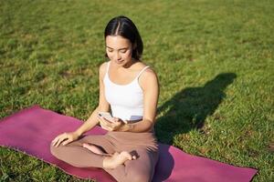 Aziatisch meisje volgt yoga opleiding app, op zoek Bij smartphone, mediteren en aan het doen opdrachten Aan vers lucht in park foto