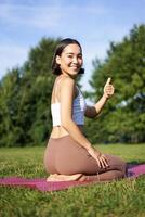 portret van Aziatisch vrouw zeggen Ja naar yoga opleiding in park, maakt duim omhoog, zittend en mediteren foto