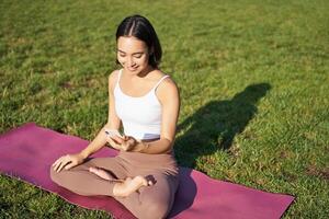 Aziatisch meisje volgt yoga opleiding app, op zoek Bij smartphone, mediteren en aan het doen opdrachten Aan vers lucht in park foto