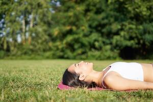 jong geschiktheid meisje aan het liegen Aan sport mat Aan gazon, ademen en mediteren in park in sportkleding foto
