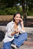portret van gelukkig jong Aziatisch vrouw pratend Aan mobiel telefoon in park, zittend Aan bank en hebben een telefoon telefoongesprek, chatten levendig foto