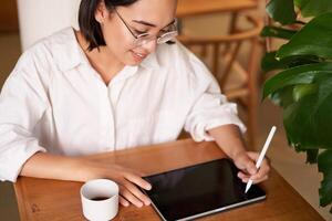 jong Aziatisch creatief meisje, zittend met digitaal tablet, tekening met grafisch pen en lachend, krabbelen, drinken koffie in cafe foto