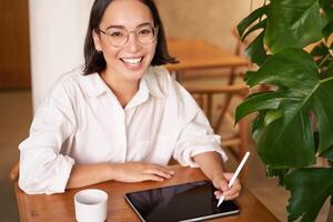 glimlachen jong vrouw Schepper, creatief meisje tekening Aan digitaal tablet met grafisch pen, drinken koffie in cafe foto