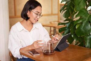 Aziatisch vrouw in bril, aan het kijken iets Aan digitaal tablet, drinken koffie in een cafe en lachend, werken van een afstand foto