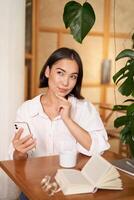 mooi jong vrouw, 25 jaren oud, denken, Holding smartphone en op zoek attent, zittend in cafe, beslissen iets foto