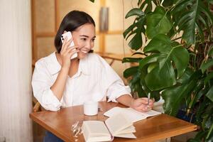 werken vrouw antwoord telefoon telefoontje in cafe, schrijven omlaag, maken aantekeningen terwijl hebben gesprek Aan telefoon foto