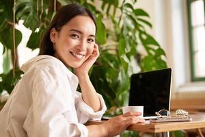 modieus jong vrouw zittend in cafe en glimlachen Bij camera, drinken koffie en gebruik makend van laptop, werken van een afstand, aan het studeren of browsen internet foto