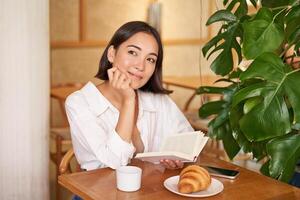 dromerig jong glimlachen Aziatisch vrouw lezing boek, zittend in cafe, aan het eten croissant en drinken koffie in knus interieur foto