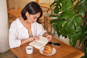 mooi jong Aziatisch vrouw zittend in cafe met een boek, aan het eten croissant en lezing, drinken kop van koffie foto