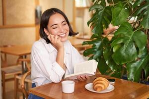 romantisch Aziatisch vrouw zittend met boek in cafe, aan het eten croissant en drinken koffie, lezing en lachend, genieten van alleen tijd foto