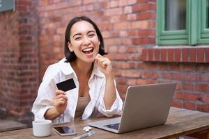 gelukkig jong Aziatisch vrouw zittend in de buurt laptop, Holding credit kaart, betalen rekeningen, boodschappen doen online contactloos, glimlachen Bij camera foto