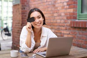 gelukkig jong Aziatisch vrouw, zittend met laptop en tonen credit kaart, betalen rekeningen online, boodschappen doen in internetten, zittend in cafe met koffie foto