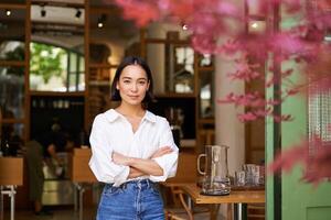 portret van jong zakenvrouw in haar eigen cafe, manager staand in de buurt Ingang en uitnodigend jij, poseren in wit duidelijk overhemd en jeans foto