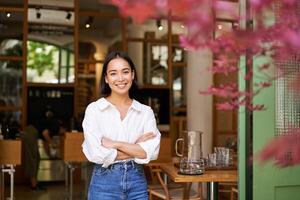 portret van jong zakenvrouw in haar eigen cafe, manager staand in de buurt Ingang en uitnodigend jij, poseren in wit duidelijk overhemd en jeans foto