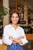 portret van jong vrouw ondernemer, cafe baasje, staand Bij de deur Ingang en lachend, op zoek zelfverzekerd foto