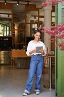 verticaal schot van glimlachen Aziatisch zakenvrouw, serveerster staand in voorkant van cafe Ingang, uitnodigend gasten in restaurant met tablet foto