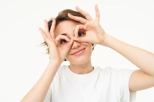 dichtbij omhoog van zorgeloos, schattig jong vrouw toont vinger bril over- haar ogen en lachend, geïsoleerd over- wit achtergrond foto