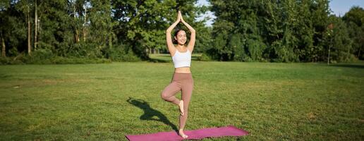 opmerkzaamheid en welzijn. jong vrouw aan het doen yoga, staand Aan mat in park, maken asana, mediteren Aan vers lucht, stedelijk leven concept foto