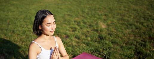 verticaal schot van jong Aziatisch vrouw aan het doen yoga, praktijk opmerkzaamheid, glimlachen en op zoek ontspannen, zittend Aan rubber mat in park foto