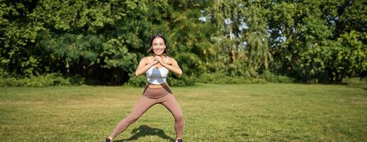 welzijn en sport complex. jong Aziatisch vrouw uitrekken, aan het doen squats en training Aan vers lucht, glimlachen verheugd foto