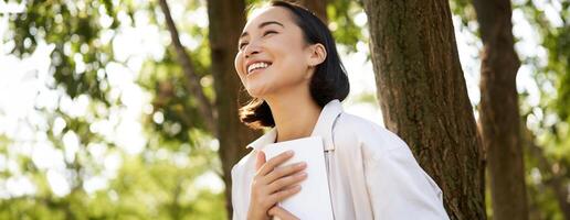 romantisch glimlachen meisje lezing boek in park of voorspel, zittend onder boom schaduw Aan zonnig dag, ontspannende Aan vers lucht omringd door natuur foto