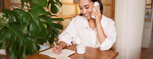 werken vrouw antwoord telefoon telefoontje in cafe, schrijven omlaag, maken aantekeningen terwijl hebben gesprek Aan telefoon foto