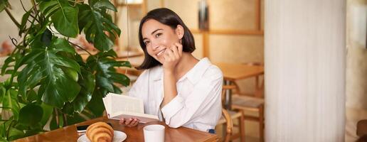 romantisch Aziatisch vrouw zittend met boek in cafe, aan het eten croissant en drinken koffie, lezing en lachend, genieten van alleen tijd foto