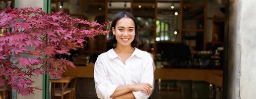 jong Aziatisch vrouw, cafe eigenaar of manager, staand zelfverzekerd met armen gekruiste Aan handen, glimlachen en op zoek Bij camera foto