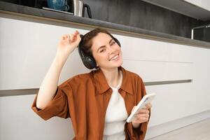modern gelukkig vrouw, zit Aan keuken verdieping Bij huis, luistert muziek- in koptelefoon, houdt smartphone, looks positief en blij foto