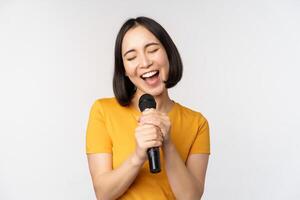 gelukkig Aziatisch meisje het zingen en hebben plezier, Holding microfoon Bij karaoke, staand in geel t-shirt tegen wit achtergrond foto
