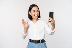 beeld van elegant modern Aziatisch vrouw golvend hand- Bij smartphone voorkant camera, video chatten, chatten met persoon Aan mobiel telefoon sollicitatie, wit achtergrond foto