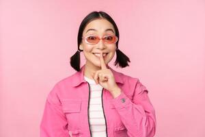 modern Koreaans meisje in elegant voorjaar kleding, zonnebril, tonen zwijgen, stil teken, druk op vinger naar lippen, taboe gebaar, staand over- roze achtergrond foto