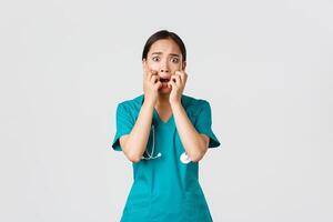 covid19, gezondheidszorg arbeiders en voorkomen virus concept. bang onzeker Aziatisch vrouw dokter, verpleegster in scrubs begint naar paniek van angst, bijten vingernagels en op zoek geschokt, wit achtergrond foto