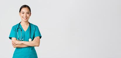 covid19, gezondheidszorg arbeiders, pandemisch concept. portret van zelfverzekerd lachend, aantrekkelijk Aziatisch vrouw verpleegster in schrobben, met stethoscoop, kruis armen borst en op zoek Bij camera, wit achtergrond foto