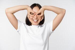 grappig jong Aziatisch vrouw, Koreaans meisje maken ogen bril gebaar, op zoek gelukkig Bij camera, staand over- wit achtergrond foto