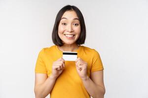 portret van mooi Koreaans meisje Holding credit kaart, aanbevelen bank onderhoud, staand in geel t-shirt over- wit achtergrond foto