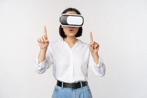 beeld van Aziatisch vrouw in vr bril richten vingers omhoog, op zoek versteld staan en verrast. meisje gebruik makend van virtueel realiteit koptelefoon, concept van toekomst en communicatie, wit achtergrond foto