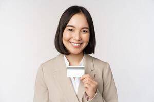 glimlachen kantoor klerk, Aziatisch zakelijke vrouw tonen credit kaart, staand over- wit achtergrond in beige pak foto