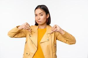 beeld van ten volle teleurgesteld Aziatisch vrouw tonen duimen omlaag, beven hoofd niet tevreden, afkeer iets, staand over- wit achtergrond foto