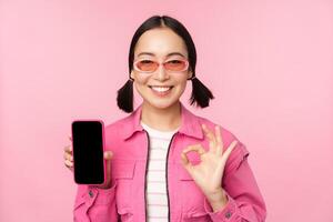 enthousiast jong Aziatisch vrouw tonen Oke, OK teken, glimlachen verheugd, mobiel telefoon scherm, smartphone sollicitatie, staand over- roze achtergrond foto