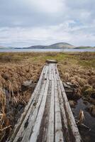 een oud houten brug gaan Rechtdoor naar de water door moerassig bodem met droog gras. donker en zwaarlijvig wolken in de lucht, verder van de meer stijgen klein bergen. foto