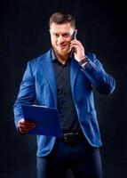 gelukkig geslaagd jong zakenman in blauw pak pratend Aan mobiel telefoon over- zwart achtergrond foto