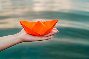 vrouw hand- houdt oranje origami boot over- de schoon blauw water. vrouw Holding handgemaakt origami boot Aan rivier- achtergrond. detailopname foto